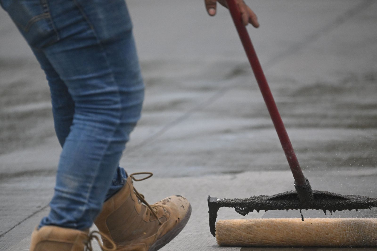 Worker in boots rolling concrete sealer across a slab with a long-handled roller