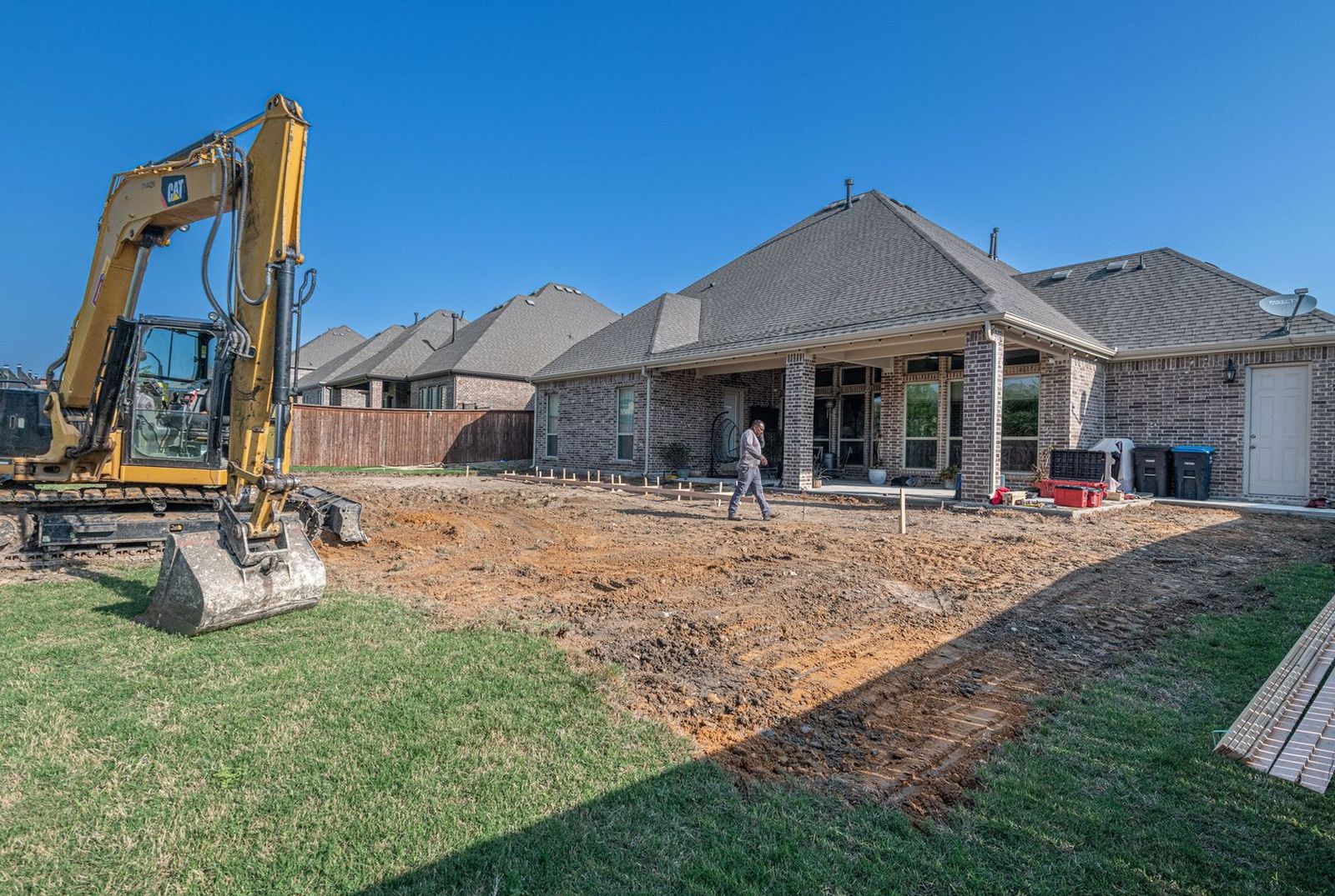 Excavator and string lines laying out a new backyard patio behind a brick house
