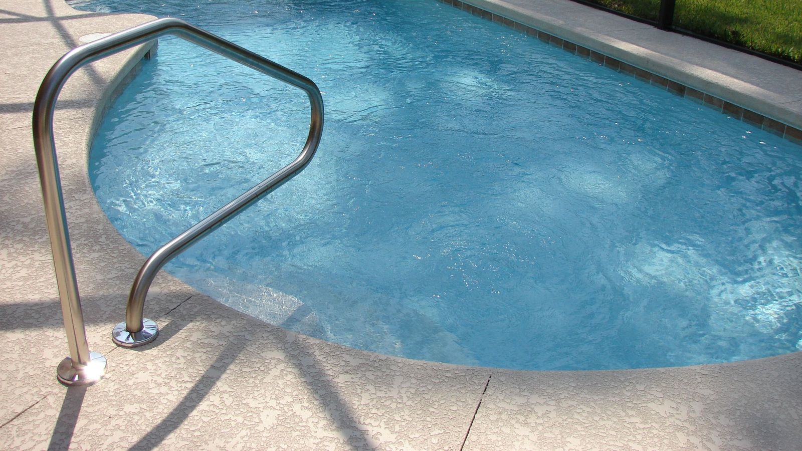 Light textured concrete pool deck curving around a backyard pool with a stainless handrail